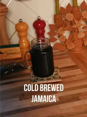 A glass mug filled with dark liquid sits on a small patterned coaster, against a backdrop of a kitchen countertop. Two decorative pepper grinders and a display of sprigs are in the back. The text "COLD BREWED JAMAICA" is overlaid at the bottom.