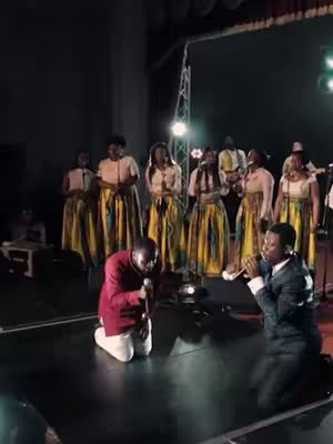 A live performance on stage with a group of choir members and two lead singers. The choir, composed of both men and women, are dressed in matching attire. The color scheme is dominated by whites and vibrant yellows. The lighting creates a dramatic atmosphere with spotlights.