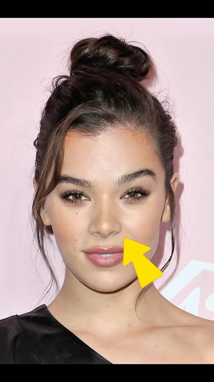 A close-up shot of a woman with brown hair pulled into a bun. Her eyes are brown, and she's smiling slightly. A yellow arrow points to her cheek. The background is a light pink.