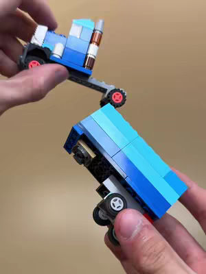 A close-up shot shows two hands holding small Lego truck models. The foreground truck is tilted towards the viewer, showing the underside and front wheels. The background truck is held higher, displaying the cab and a portion of the trailer. The colors of the Legos are mostly blue, with some white and brown accents. The background is a neutral tan.