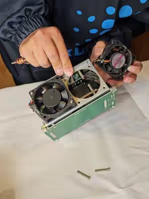 A person is holding an open GPU with fans and a screwdriver, with tools and parts visible on a white surface. The person is wearing a dark jacket. A second fan is being held in the other hand. Small screws are at the bottom.