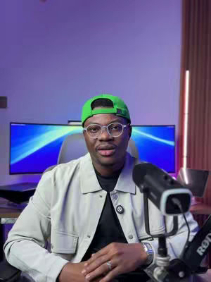 A man wearing glasses and a green hat sits in front of a computer screen with blue and purple graphics. He is wearing a light jacket over a black shirt, and a microphone is positioned near him. The background is a gradient of purple hues.