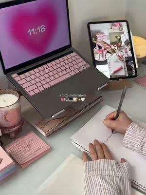 An overhead shot presents a workspace with a laptop displaying a pink heart graphic on its screen. A tablet is propped up next to it, showing a grid of images. There's a notebook with handwriting on the right, a candle, and various stationary items like sticky notes and pens scattered around the desk. The color scheme is light, with pink and white tones dominating.