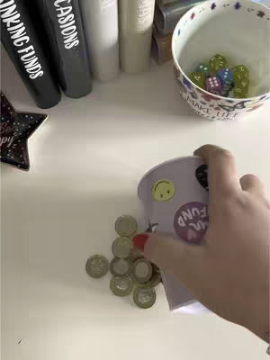 A hand holding a small container, pouring out a small pile of pound coins onto a table. The background shows various items, including books and a bowl with dice in it.