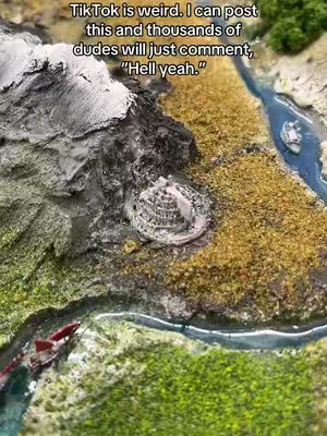 An aerial view of a detailed miniature landscape. The terrain is constructed with varying textures and colors, creating a realistic depiction of mountainous regions and waterways. A large, textured mountain dominates the left side, while a detailed model of a castle-like structure sits centrally. A winding river with a small boat flows through the landscape. The overall impression is of a meticulously crafted fantasy world.