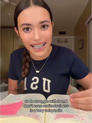 A young woman with dark hair in a braid is centered in a well-lit indoor space. She wears a navy blue t-shirt with the word "USA" in white block letters. She is smiling with braces visible, and a necklace is around her neck. She is looking directly at the camera, and holding the pages of a book open in front of her. The text overlay is positioned below her, indicating an interior setting.