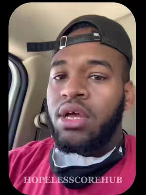 A man wearing a black baseball cap and a red shirt is sitting in a car. The man's facial expression is one of curiosity, with his mouth slightly open and a beard framing his face. He holds a black mask under his chin.
