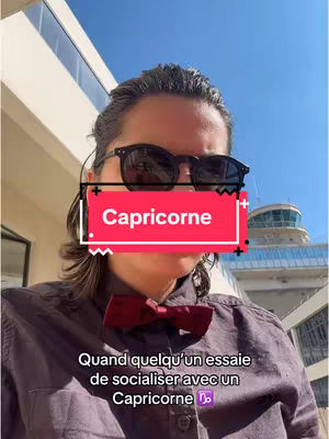 A selfie taken against a clear blue sky, featuring a person wearing black sunglasses and a burgundy bow tie. A red box with the word "Capricorne" covers part of the person's face. The background reveals a building with a control tower.