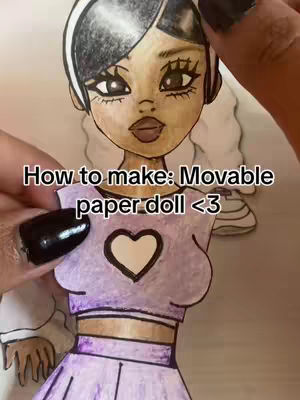 A close-up shot shows a partially completed paper doll, held by a hand with black nail polish. The doll features a stylized anime-inspired design with black and white hair, large eyes, and a soft makeup look. The doll wears a lavender top with a white heart and a matching skirt. Text overlayed reads 'How to make: Movable paper doll <3'. The background is a crafting scene suggesting a DIY tutorial.