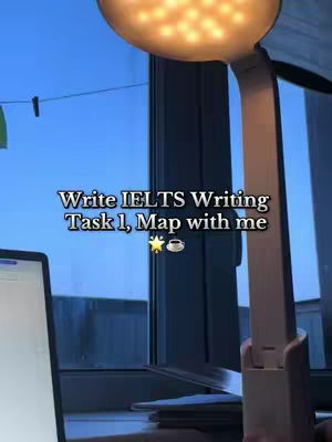 The cover image is an indoor shot. An adjustable desk lamp with a round, glowing light source is prominent on the right side. A laptop with a lit screen is partially visible on the left. The background shows a window with a view of an outdoor scene with light blue sky and a black clothespin. Text is overlaid to promote IELTS study content.