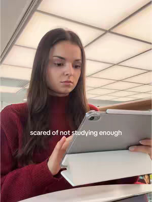 A young woman is looking down at a tablet device. She has long brown hair and is wearing a burgundy turtleneck sweater. The background is out of focus but appears to be an interior setting, perhaps an office. The overlaying text reads 'scared of not studying enough'.