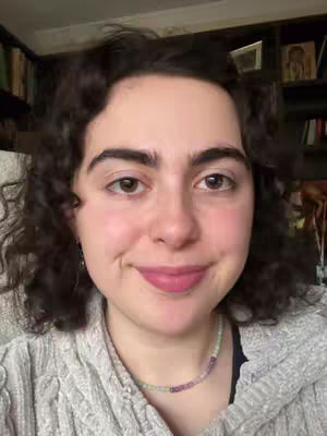 A woman with curly dark hair and fair skin is centered in a selfie-style shot. She has brown eyes, lightly defined eyebrows, and wears delicate earrings. Her lips are colored with a pale pink lipstick. She's dressed in a gray, textured sweater, and a beaded necklace with pink and green beads is visible. The background is subtly blurred but includes a bookshelf and a hint of a framed picture. The overall lighting is soft and even, contributing to a warm, inviting feel.