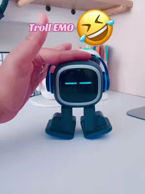 The cover image shows a small, dark-colored robot toy with a square screen and headphones sitting on a white surface. A hand with a ring is placed on top of the robot. Overlayed text says 'Troll EMO'. A laughing emoji is also placed on the image.