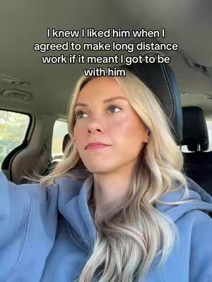 A woman with long blonde hair is sitting in the driver's seat of a car. The image is slightly angled, capturing her looking off to the side. The background shows the car's interior.