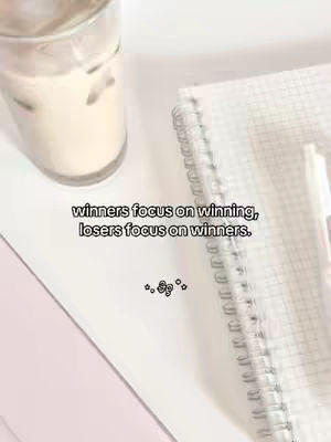 The cover image displays a close-up shot of a white, organized surface. A clear glass with a light-colored beverage and ice cubes sits in the upper left corner. To the right, a white spiral-bound notebook with grid-lined paper lies open. A pen is placed on the notebook. A handwritten quote in a simple black font is centered across the image that reads, 'winners focus on winning, losers focus on winners.' Below the quote is some decorative elements of the same color. The overall aesthetic uses a clean, minimalist design, with a soft, neutral color palette dominated by whites and off-whites. The composition emphasizes the message by using a centered layout with balanced elements.