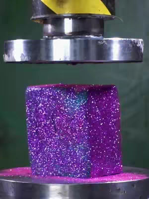 The image shows a close-up of a purple, glitter-covered cube being crushed by a large, metal press or machine. The cube is sitting on a metal base, and the top portion of the press is coming down from above. The color palette is dominated by shades of purple, pink, silver, and gray.