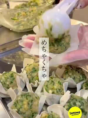 An overhead shot of a person arranging a filled dumpling into a paper cup, with more dumplings in trays below. The food is green and white indicating ingredients like green vegetables, in a home-cooking setting. Text overlays on the image feature Japanese text and the Lemon8 logo.