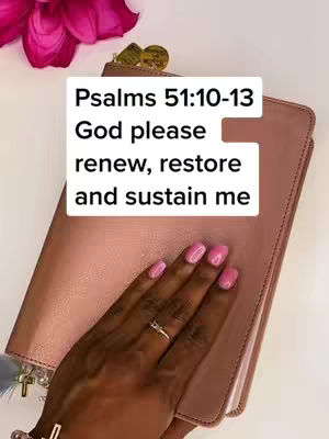 A close-up shot focuses on a woman's hand resting on a pink Bible. The hand, adorned with pink nail polish and a ring, holds the Bible open. A white text overlay reading 'Psalms 51:10-13 God please renew, restore and sustain me' is positioned in the center-left of the image. The background is a light surface, accented by a pink flower in the top right corner.