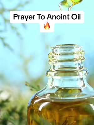A close-up shot of a clear glass bottle filled with amber-colored liquid, presumably oil. The top of the bottle is in focus, with drops of oil being poured in from above. The background is a soft blur of green and blue, possibly foliage and sky.