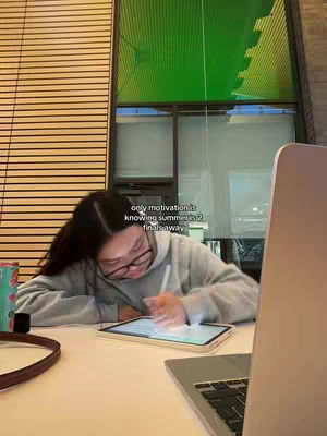A person, wearing glasses and a hoodie, is hunched over a table in a library setting. They are using a tablet with a stylus and a laptop is partially visible in the right corner. Text overlayed says: "only motivation is knowing summer is 2 finals away."