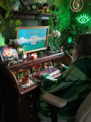 A person sits at a rolltop desk, illuminated by green lighting. A monitor displays a game screen, with a keyboard in front and various decor items like candles and greenery surrounding the setup. The overall aesthetic is dark cottagecore with a cozy, maximalist vibe.