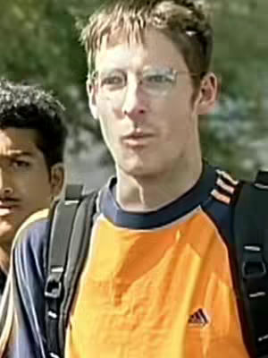 A medium shot shows a man wearing glasses, an orange and blue shirt, and a backpack. Another person is partially visible in the background on the left side.