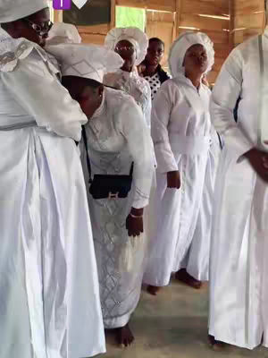Several people dressed in white religious garments, standing in what appears to be a wooden-walled room. The individuals are of varying heights and ages. Focus is on a person in the foreground who appears to be bowing down. There is some natural light in the room, and the background is slightly blurred.