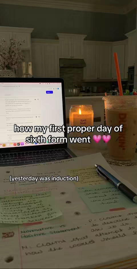 A photo of a desk setup shows a laptop displaying text, with a Dunkin' iced coffee, a lit candle, and handwritten notes. The notes and laptop suggest a study environment.