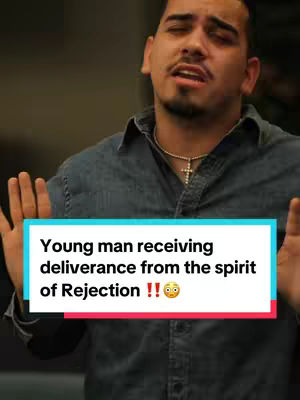 A young man is featured, mid-shot, with his arms up and eyes closed, potentially in a moment of prayer or reflection. He wears a denim shirt and a gold chain with a cross. Overlaying him is a rectangular text box with the words 'Young man receiving deliverance from the spirit of Rejection !!' in white font against a turquoise and red backdrop.