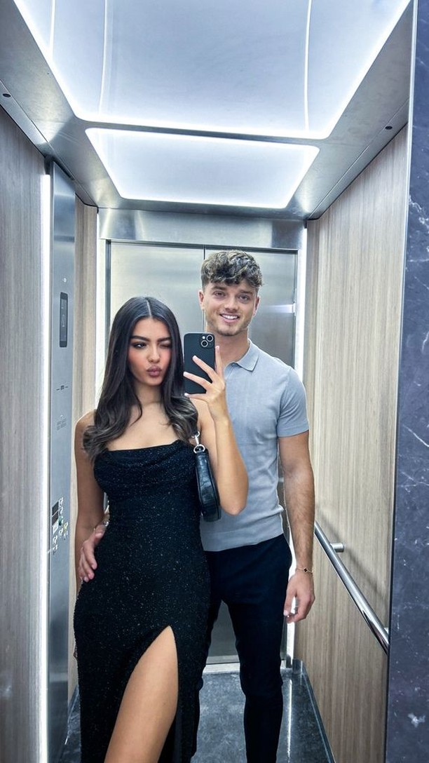 A couple takes a mirror selfie in an elevator. The woman is in a black dress with a thigh-high slit. The man is behind her with a gray polo and black pants, smiling while holding the phone. The interior of the elevator has wood paneling and bright overhead lighting.