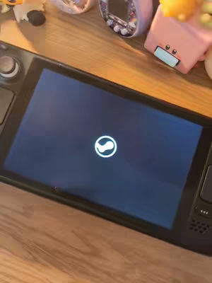 A Steam Deck device is displayed on a wooden surface. The screen shows a dark blue background with a white Steam logo in the center. Various small toys and accessories are scattered around the Steam Deck, adding a playful touch to the composition.