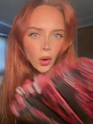 Close-up shot of a young woman with fair skin and reddish-orange hair, her mouth is open in an expression of surprise, and a patterned garment is blurred in the foreground.