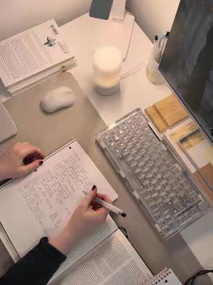 A top-down view of a well-lit desk with a notebook where someone is writing, surrounded by books, a keyboard, a mouse, and a lamp.