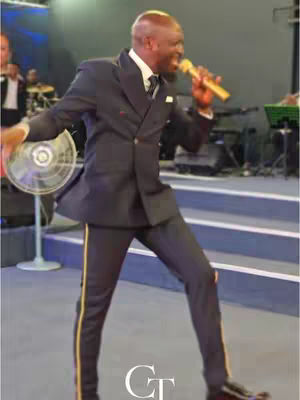 The image shows a man in a formal dark suit with a microphone, appearing to be giving a rousing speech or performance on a stage. The man is animatedly gesturing with his arms, and a fan is behind him. The lighting suggests a stage environment with other musicians and equipment visible in the background.