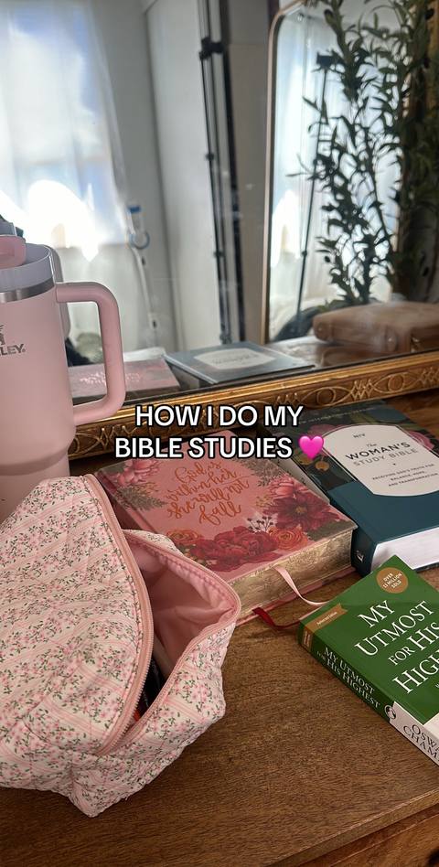 The image shows a well-lit interior scene, likely a desk or table, with various items suggesting a focus on Bible study. Several books are prominently displayed, including what appears to be a 'Woman's Study Bible' and other volumes with floral covers. A pink insulated tumbler sits on the left, and a floral-patterned makeup bag is partially open in the foreground. The overall composition is warm and inviting, with soft lighting and a focus on the books and study materials.