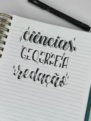 An overhead shot of a notebook with lined paper. The notebook is open to a page displaying several lines of black brush lettering that feature the words "ciências", "geografia", and "redação", with decorative starbursts. A black pen is placed diagonally in the upper right corner. The background is a plain white surface.