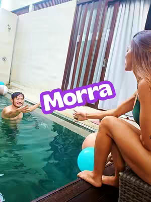 A woman sits poolside with one leg crossed, smiling and gesturing towards a man in the pool. The background includes a pool and a natural setting.