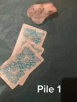 The image features three tarot cards fanned out, a crystal rock, and text overlayed on a slightly textured teal surface. The cards are face up, showing floral designs in blue and white. The crystal is in the upper right, and the text "Pile 1" is at the image's bottom right.