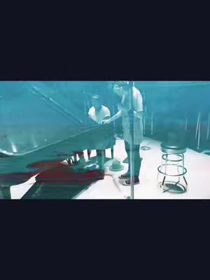 The cover image presents a split-screen scene featuring a lower portion with two people standing next to a large piano, while the upper portion is a solid, dark blue. The focus is on the interior space with bluish lighting. The people wear casual attire. A metallic stool and a hat are present.