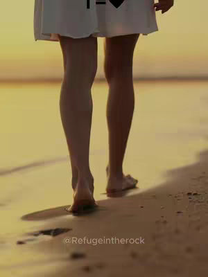 A low-angle shot from behind of a woman with bare feet walking away from the camera on a sandy beach. The sun is setting in the background, creating a warm, golden hue. The woman is wearing a white dress. Her legs are visible, and she leaves footprints in the wet sand. The overall tone is peaceful and serene.