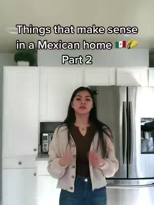 A woman stands in a kitchen setting in front of white cabinets and a stainless steel refrigerator. The text overlay reads 'Things that make sense in a Mexican home Part 2'. She is wearing a light jacket, a dark top and jeans.