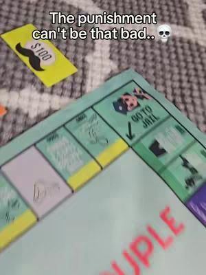 The image shows a tabletop scene focused on a board game. In the foreground, a section of the game board features the word "COUPLE" in red. A yellow card with a stylized black mustache and the text "$100" is positioned in the upper left. To the right, a portion of the board game includes the "GO TO JAIL" square. The background suggests a patterned surface. The overall shot is angled slightly.