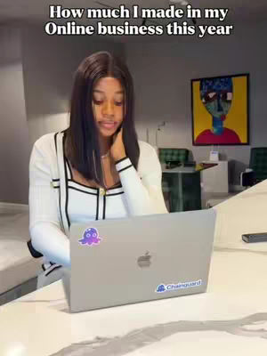 A woman is sitting in front of a laptop with stickers on it, with a painting on the wall behind her. The background is blurred, but it appears to be an office or home setting.