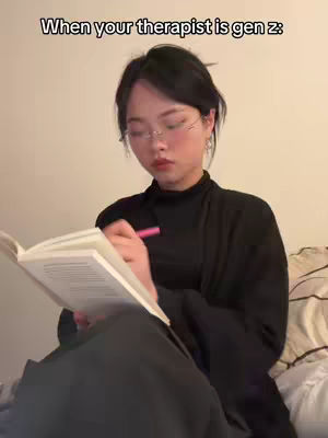 A young woman wearing glasses and a black turtleneck is seated on a bed, holding a notebook open. She has dark hair and is writing with a pink pen. The background is a plain cream-colored wall with some pillows visible behind her. The image has a calm, personal feel.