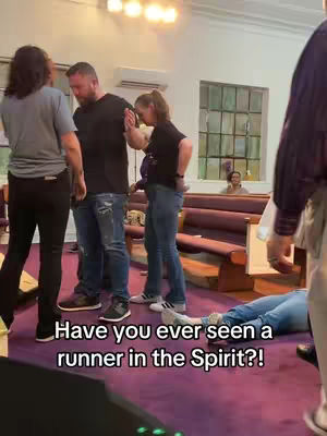 A group of people in a church setting, with individuals standing and one person lying on the ground. The floor is covered in a purple carpet. Text overlayed on the bottom.