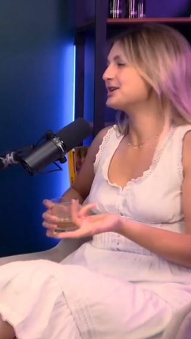 The image shows a woman in profile, seated with a microphone in front of her. She is wearing a sleeveless white dress. Her light blonde hair is styled with a side part, and she has a necklace. She is holding a glass and gesturing as if speaking, set against a blurry blue background with a dark bookshelf and a microphone in the foreground. The lighting appears to be soft.