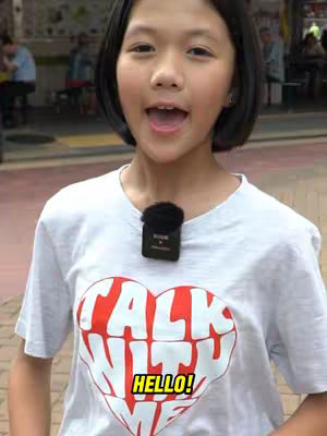 A young Asian girl with short black hair stands outside, smiling with an open mouth, wearing a white t-shirt with a red heart-shaped design. A microphone is clipped to her shirt. The setting appears to be outdoors with a blurry background.