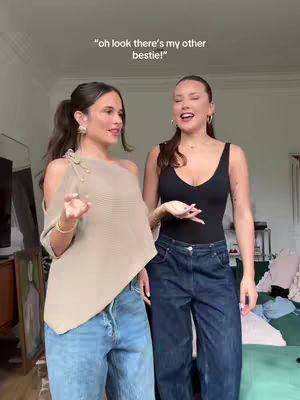 Two young women are standing indoors, likely in a home setting. The woman on the left is wearing a beige, asymmetrical top and light blue jeans. The woman the on right is clad in a black tank top and dark blue jeans. The backdrop reveals a soft-colored wall, a mirror, and parts of furniture suggesting a casual, lived-in space.