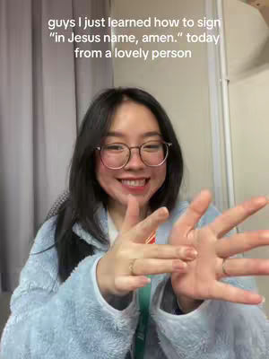 A woman with dark hair and glasses smiles at the camera while demonstrating sign language with her hands. Text on the image indicates that she is learning to sign "in Jesus name, amen."