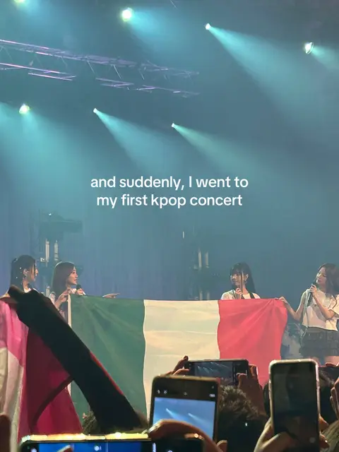 A live concert scene, shot from the perspective of the audience. Four female performers are on stage, each holding up a section of an Italian flag. The crowd is visible in the foreground with many arms holding up phones to record or take pictures. The stage is lit with vibrant lights. The text overlay says, "and suddenly, I went to my first kpop concert".